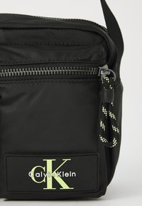 Black nylon crossbody bag with a silver zipper, prominent logo patch in green and white, and textured black strap with yellow accents.
