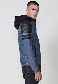 Uomo con giacca blu, spalle imbottite nere e cappuccio, in piedi di lato e che guarda oltre la spalla su uno sfondo semplice.