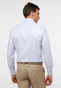 Camicia azzurro chiaro a maniche lunghe con finitura testurizzata e polsini con bottoni, abbinata a una cintura tessuta beige e pantaloni chiari.