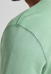 Tela de camiseta de algodón verde claro, textura suave con un escote redondeado y detalle en la costura del hombro, exhibiendo un diseño casual.