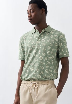 Νεαρός άνδρας που φοράει ένα πουκάμισο πόλο πράσινου χρώματος με λευκά σχέδια κοχυλιών και μπεζ παντελόνι με κορδόνι, κοιτώντας προς τα κάτω αριστερά του.