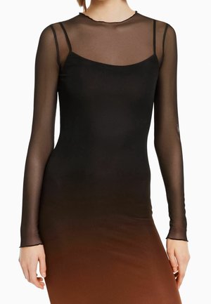 Vrouw draagt een zwarte mouwloze jurk, gelaagd onder een doorschijnende zwarte mesh top met lange mouwen en een ronde hals.