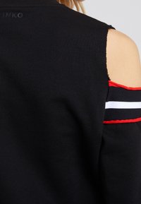 Sweatshirt noir avec un design à épaule dénudée, présentant un accent rayé rouge et blanc le long de la manche. Tissu textur é et bords bruts.