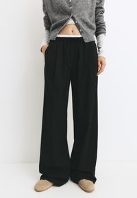 Čierne nohavice s širokými nohavicami a voľným strihom, s elastickým pásom a mäkkou tkaninou. Spárované so sivým cardiganom a béžovými topánkami.