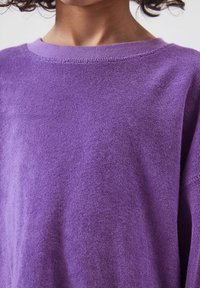 Gros plan sur une personne portant un pull violet texturé à col rond, montrant l'encolure et le haut du torse.