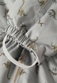 Tela con estampado de dinosaurios en gris y con una textura suave. PresentaGathering elástico y un bucle de plástico transparente para abrochar.