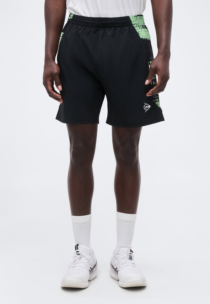 Μαύρα αθλητικά σορτς με πράσινα πλαϊνά πάνελ με μοτίβο, φορεμένα από ένα άτομο που στέκεται, με λευκές κάλτσες και λευκά αθλητικά παπούτσια, σε απλό φόντο.