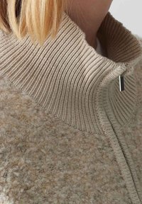 Beige fleece jacket featuring a ribbed high collar, textured body, and a side zipper with a small, rectangular pull tab.