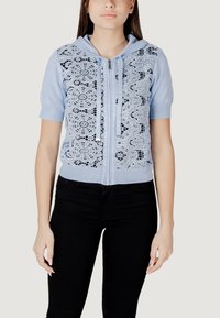 Sudadera de cremallera azul claro con mangas cortas y superposición de encaje con patrones florales; incluye cordones grises y se combina con pantalones negros.