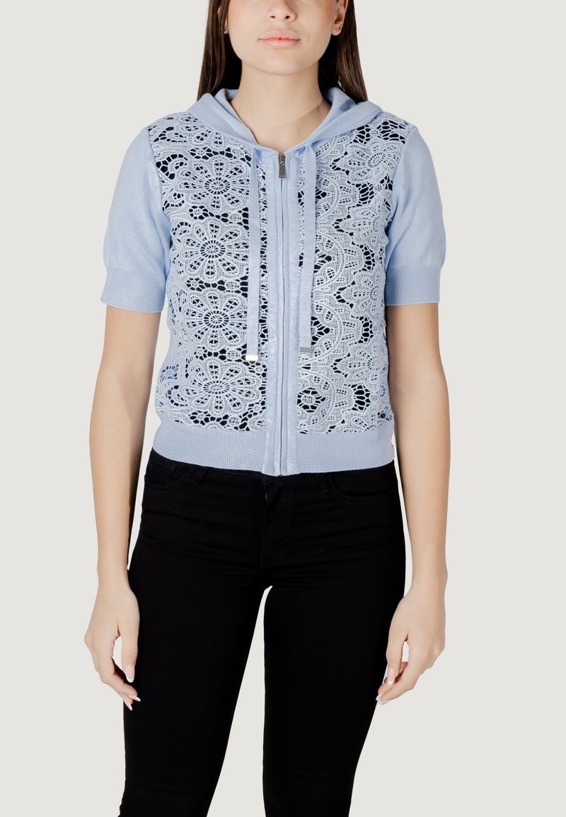 Sudadera de cremallera azul claro con mangas cortas y superposición de encaje con patrones florales; incluye cordones grises y se combina con pantalones negros.