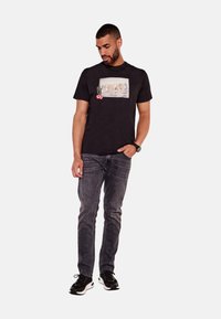 Camiseta de algodón negra con un estampado gráfico que incluye la palabra "GUESS", un cactus y acentos florales. Combinada con jeans grises y zapatillas negras.