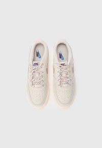 Par športnih copat Nike v krem barvi z večbarvnimi vezalkami in rjavimi znaki swoosh, pogledano od zgoraj na belem ozadju.