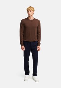 Maglione marrone a maniche lunghe, colletto rotondo, abbinato a pantaloni blu navy e sneakers bianche. Il tessuto sembra essere testurizzato e leggero.