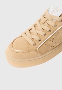 Beige Wildleder-Sneaker mit runder Zehenpartie, flachen Schnürsenkeln, goldenen Akzenten und eingeprägtem Markenlogo. Strukturierte Oberseite und Gummisohle mit glatter Oberfläche.