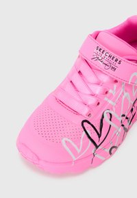 Scarpa sportiva rosa con superficie testurizzata, perforazioni, lacci e una fascia; presenta design grafici a forma di cuore in bianco e nero.