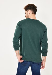 Langärmliges grünfarbenes Sweatshirt mit Rundhalsausschnitt, strukturiertes Material, bequeme Passform, hochgekrempelte Ärmel, kombiniert mit dunkelblauen Jeans, von hinten dargestellt.