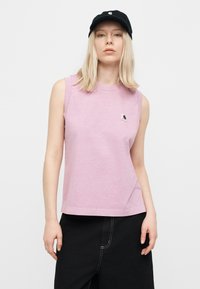 Γυναίκα με ξανθά μαλλιά που φοράει μαύρο καπέλο, αμάνικο ανοιχτό μωβ τοπ με μικρό λογότυπο και μαύρο παντελόνι σε λευκό φόντο.