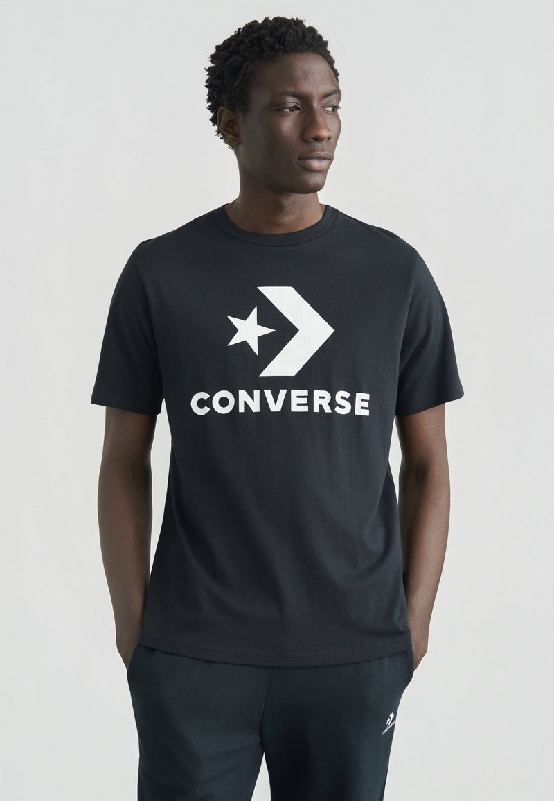 Μαύρο βαμβακερό μπλουζάκι με μεγάλο λευκό λογότυπο "CONVERSE", σχέδιο αστέρι και βέλος στο μπροστινό μέρος, με στρογγυλή λαιμόκοψη και κοντά μανίκια.