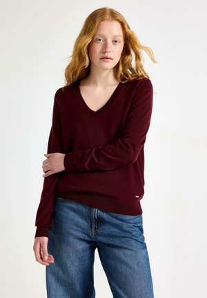 Ung kvinde med bølget rødt hår iført en burgundy V-hals sweater og blå jeans, stående med den ene arm krydset over kroppen.