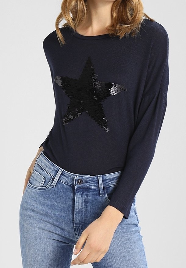 Femme portant un haut bleu marine à manches longues avec une grande étoile noire en sequins sur la poitrine et un jean taille haute bleu clair.