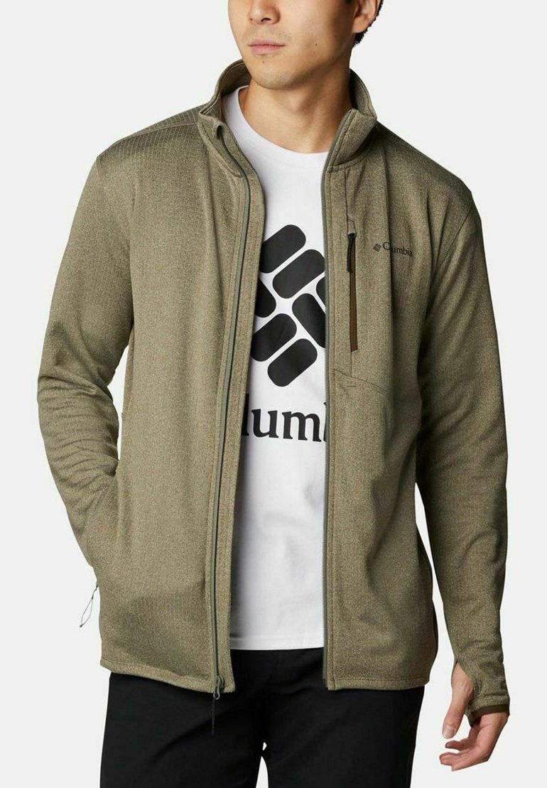 Uomo che indossa una giacca verde oliva con zip sopra una T-shirt bianca con un logo geometrico nero e pantaloni neri, posando con le mani nelle tasche.