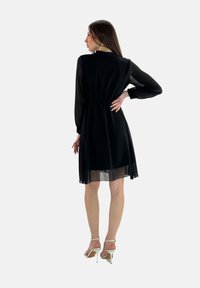 Abito di chiffon nero con maniche lunghe in semitrasparente e vita arricciata. Lunghezza a metà polpaccio con una morbida caduta e una vestibilità fluida. Indossato con tacchi bianchi con cinturini.