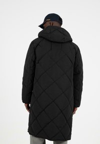 Černý prošívaný dlouhý kabát s kapucí, se vzorem diamantů a zaobleným lemem. Nositý přes šedé kalhoty, s tmavou čepicí.