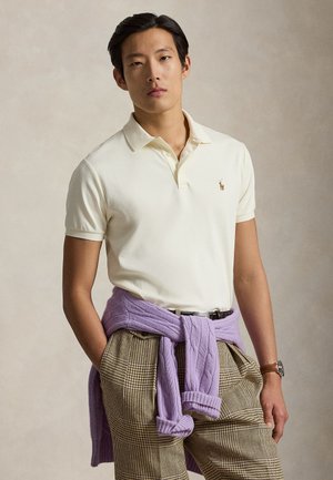 Mees, kes kannab kreemjat polosärki, ruudulisi pükse ja lavendlivärvi kampsunit, mis on vöökohale seotud, seisab käed taskutes ühevärvilise tausta ees.