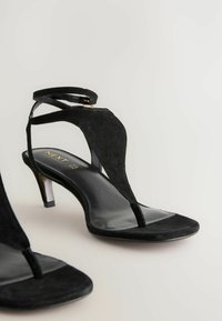 Sandali con tacco alto in suede nero, design con T-strap e cinturino alla caviglia. Presentano una soletta nera lucida e un accento metallico sul tacco.