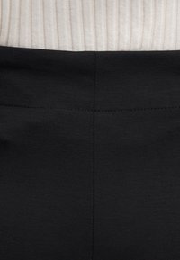 Gros plan sur un tissu noir avec un détail de couture centrale, le haut côtelé beige clair du porteur visible partiellement en haut.