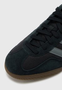 Čierna semišová a sieťovaná teniska s tmavými šnúrkami, so sivým pruhom a textúrovanou gumovou podrážkou, zobrazená zhora.