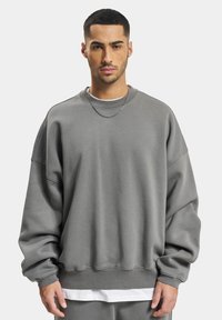 Grå oversized sweatshirt lavet af tekstureret stof, med brede ærmer, en rund hals og en ribbet kant, lagdelt over en hvid top.