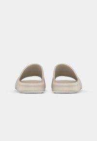 Beige slip-on sandals with open back and sides, smooth texture, rounded design, and flat sole. Minimalist style with no visible hardware.