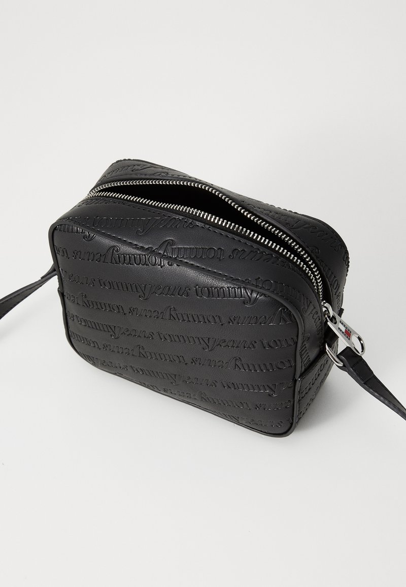 Bolsa de nailon texturizado negro con un patrón emblema "Tommy Jeans", cierre de cremallera y una correa desmontable. Forma rectangular con bordes suaves.