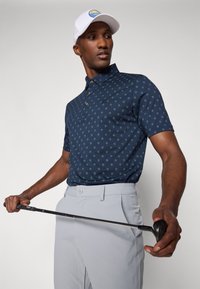 Polo navy con pequeño patrón floral verde, con placket de dos botones, usado con pantalones gris claro mientras sostiene un palo de golf. Gorro blanco en la cabeza.