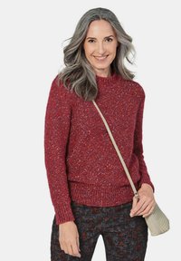 Handgestrickter roter Pullover mit strukturiertem Finish, rundem Ausschnitt und geripptem Saum, kombiniert mit dunklen gemusterten Hosen und einer beigen Umhängetasche.