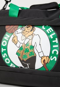 Juodas sportinis krepšys su spalvingu Bostono "Celtics" logotipu, kuriame vaizduojamas leprachanas, žali akcentai ir krepšinio kamuolys.