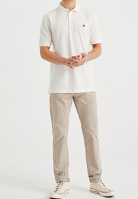 Biała koszulka polo z krótkim rękawem i małym zielonym logo, w połączeniu z jasnobeżowymi spodniami w prążki i kremowymi sneakersami.