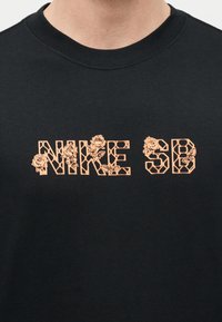 Μαύρο μπλουζάκι με στρογγυλή λαιμόκοψη και το κείμενο «NIKE SB» σε ροδακινί γράμματα με περίγραμμα, διακοσμημένα με λεπτομερείς εικονογραφήσεις ροδακινί τριαντάφυλλων.