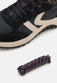 Schwarze und graue Sneaker aus Wildleder und synthetischen Materialien, mit einem beigen Akzentstreifen; eine gedrehte marineblaue Schnürsenkel mit roten Streifen darunter.