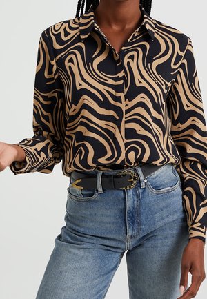 Kvinde iført en sort langærmet skjorte med beige abstrakte hvirvler, stoppet ind i blå jeans med et sort bælte med en guldfarvet spænde.