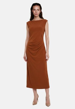 Donna che indossa un vestito midi senza maniche color ruggine con arricciature laterali e tacchi alti beige, in piedi davanti a uno sfondo bianco uniforme.