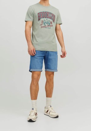 T-shirt en coton vert clair avec le texte imprimé "BROOKLYN", shorts en denim bleu et baskets beiges avec des accents bordeaux et des chaussettes blanches.