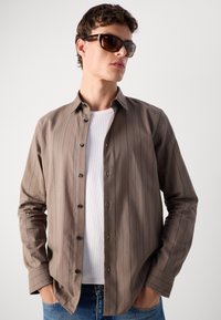 Camicia a righe marroni con bottoni neri, indossata sopra una t-shirt bianca, abbinata a occhiali da sole. Il tessuto appare liscio e con una texture morbida.