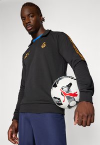 Sort langærmet polo med gyldne logo accenter og en blå krave, parret med marineblå shorts. Holder en sort og hvid fodbold med rødt logo.