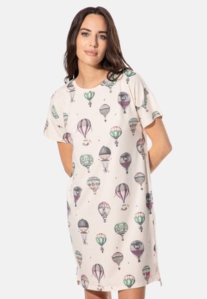 Frau in einem cremefarbenen Kleid mit bunten Heißluftballon-Prints, die mit verschränkten Händen vor schlichtem Hintergrund steht.