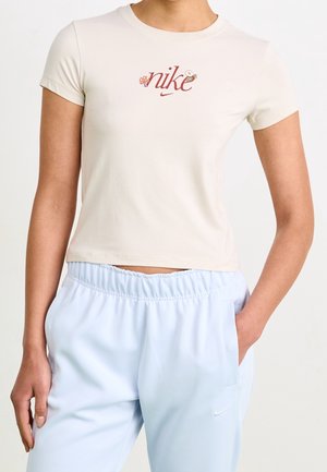 Kobieta w beżowej krótkiej koszulce Nike z kwiatowymi detalami oraz jasnoniebieskich spodniach dresowych Nike z elastycznym pasem i bocznymi kieszeniami.
