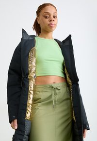 Ung kvinde med fletning, iført en sort frakke med guldforing, en lysegrøn cropped tanktop og en olivengrøn nederdel med snøre.