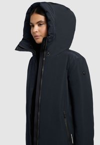 Marineblå vandtæt pufferjakke med stor hætte, lynlåslukning foran, sidelommer og struktureret stof. Har elegante sorte detaljer.