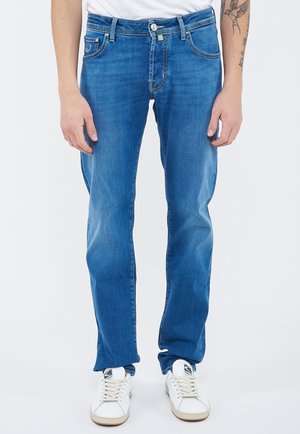 Man wearing blue straight-leg jeans and white sneakers, standing against a plain white background.